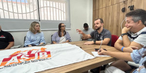 A imagem mostra uma reunião em uma sala iluminada com mesa retangular de madeira, onde oito pessoas estão sentadas, participando de uma conversa atenta. No centro da mesa, há uma bandeira estendida com o logotipo da ASJB. As expressões dos participantes revelam concentração e interesse no tema discutido. Trata-se de um encontro com aparência formal, voltado à representação ou debate de assuntos relacionados à categoria.