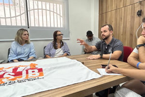 A imagem mostra uma reunião em uma sala iluminada com mesa retangular de madeira, onde oito pessoas estão sentadas, participando de uma conversa atenta. No centro da mesa, há uma bandeira estendida com o logotipo da ASJB. As expressões dos participantes revelam concentração e interesse no tema discutido. Trata-se de um encontro com aparência formal, voltado à representação ou debate de assuntos relacionados à categoria.