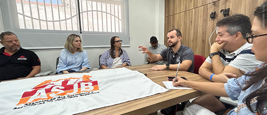 A imagem mostra uma reunião em uma sala iluminada com mesa retangular de madeira, onde oito pessoas estão sentadas, participando de uma conversa atenta. No centro da mesa, há uma bandeira estendida com o logotipo da ASJB. As expressões dos participantes revelam concentração e interesse no tema discutido. Trata-se de um encontro com aparência formal, voltado à representação ou debate de assuntos relacionados à categoria.