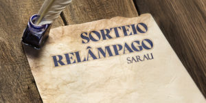 Imagem Acessível: A imagem apresenta um convite visual para um "Sorteio Relâmpago" relacionado a um sarau com participação da filiada do SINJUS-MG. Em destaque, há uma folha de papel envelhecido sobre uma mesa de madeira rústica, com um tinteiro azul-escuro e uma pena repousando sobre ela — remetendo à escrita clássica. No papel, lê-se em letras grandes: “SORTEIO RELÂMPAGO - SARAU”, seguido do tema “A tirania do gozo imediato x A última dor que me salvou”. Abaixo, há informações práticas: inscrições até o dia 8 de maio (quinta-feira), resultado no dia 9 (sexta-feira), e datas dos eventos — 10 de maio (sábado) às 20h e 11 de maio (domingo) às 19h, no Teatro da Biblioteca Pública Estadual de Minas Gerais. A marca do SINJUS-MG aparece na parte inferior da imagem. O visual retrata um evento cultural com atmosfera reflexiva e artística.