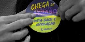 Imagem Acessível: Imagem em fundo preto, com uma fotografia em preto e branco de mãos segurando um material circular colorido. No material, lê-se “Chega de cultura do atraso” e “Data-base é obrigação!”. Conteúdo textual: Participe - SINJUS convoca categoria para Ato Público contra os atrasos do TJMG.