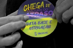 Imagem Acessível: Imagem em fundo preto, com uma fotografia em preto e branco de mãos segurando um material circular colorido. No material, lê-se “Chega de cultura do atraso” e “Data-base é obrigação!”. Conteúdo textual: Participe - SINJUS convoca categoria para Ato Público contra os atrasos do TJMG.