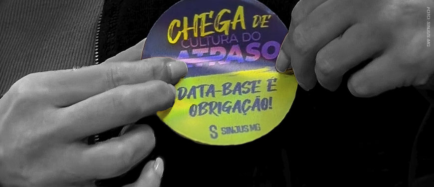 Imagem Acessível: Imagem em fundo preto, com uma fotografia em preto e branco de mãos segurando um material circular colorido. No material, lê-se “Chega de cultura do atraso” e “Data-base é obrigação!”. Conteúdo textual: Participe - SINJUS convoca categoria para Ato Público contra os atrasos do TJMG.