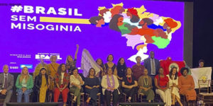 Imagem Acessível:Fotografia composta pelas representantes do Ministério das Mulheres, em cima de um palco onde se vê ao fundo a arte da campanha #BrassilsemMisoginia com o fundo lilás com o desenho do mapa do Brasil com diversas mulheres.