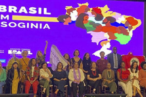 Imagem Acessível:Fotografia composta pelas representantes do Ministério das Mulheres, em cima de um palco onde se vê ao fundo a arte da campanha #BrassilsemMisoginia com o fundo lilás com o desenho do mapa do Brasil com diversas mulheres.