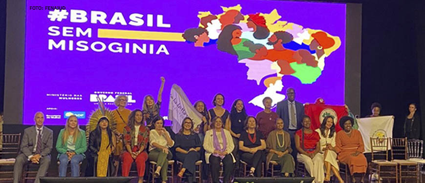 Imagem Acessível:Fotografia composta pelas representantes do Ministério das Mulheres, em cima de um palco onde se vê ao fundo a arte da campanha #BrassilsemMisoginia com o fundo lilás com o desenho do mapa do Brasil com diversas mulheres.