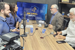Cena de estúdio de gravação de videocast com quatro pessoas sentadas ao redor de uma mesa com microfones. Um homem de camisa azul, Felipe Rodrigues (diretor de Assuntos Jurídicos do SINJUS-MG) fala enquanto os demais o escutam. Ao lado do diretor do SINJUS está a defensora pública geral, Raquel da Costa Dias, à frente deles estão o jornalista e anfritrião Orion Teixeira e o diretor da AFFEMG, Lucas Espechit. Ao fundo, há uma TV com o logotipo do programa “Se Liga, Minas!”.