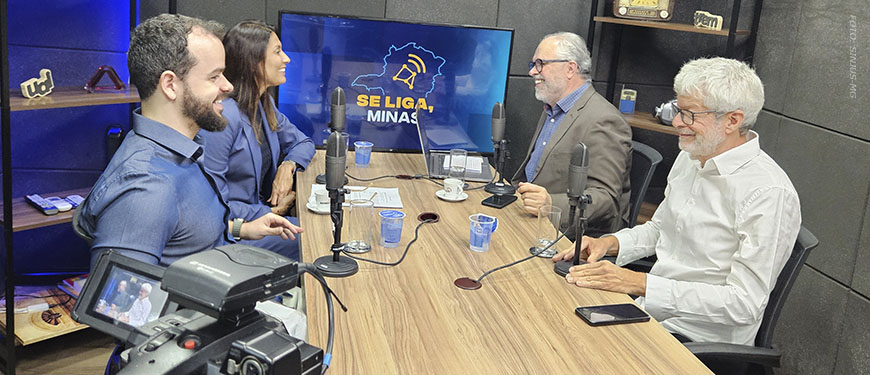 Cena de estúdio de gravação de videocast com quatro pessoas sentadas ao redor de uma mesa com microfones. Um homem de camisa azul, Felipe Rodrigues (diretor de Assuntos Jurídicos do SINJUS-MG) fala enquanto os demais o escutam. Ao lado do diretor do SINJUS está a defensora pública geral, Raquel da Costa Dias, à frente deles estão o jornalista e anfritrião Orion Teixeira e o diretor da AFFEMG, Lucas Espechit. Ao fundo, há uma TV com o logotipo do programa “Se Liga, Minas!”.