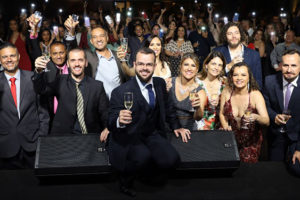 Em primeiro plano, estão diretoras, diretores e suplentes do corpo diretivo do SINJUS, juntos eles levantam um brinde. Ao fundo, há um grande número de pessoas com suas lanternas dos celulares acesas.