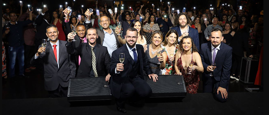 Em primeiro plano, estão diretoras, diretores e suplentes do corpo diretivo do SINJUS, juntos eles levantam um brinde. Ao fundo, há um grande número de pessoas com suas lanternas dos celulares acesas.