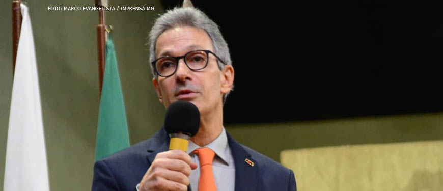 No primeiro plano está um senhor esbelto de cabelos grisalhos e curtos, discursando em um microfone sobre um púlpito. Ao fundo estão as bandeiras do Brasil e de Minas Gerais.