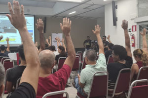 Na imagem há um auditório com diversas pessoas assistindo a uma apresentação que está no telão. De frente para a plateia está Alexandre Pires, com o braço levantado, ele é um homem branco e está vestido com uma camisa preta e calça jeans. As pessoas presentes no auditório repetem o gesto, levantando também os braços para o alto em sinal de aprovação à pauta proposta pelo Sindicato.