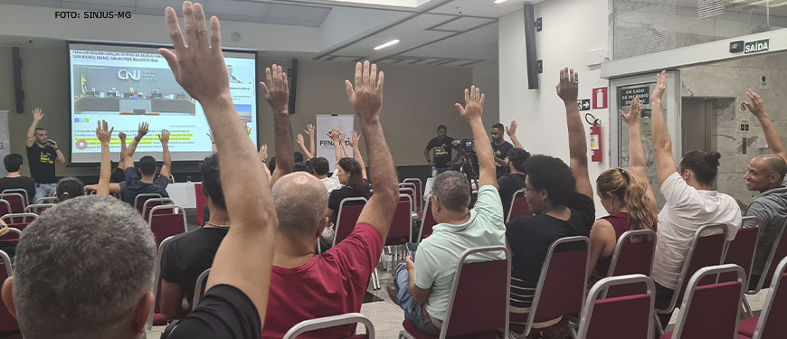 Na imagem há um auditório com diversas pessoas assistindo a uma apresentação que está no telão. De frente para a plateia está Alexandre Pires, com o braço levantado, ele é um homem branco e está vestido com uma camisa preta e calça jeans. As pessoas presentes no auditório repetem o gesto, levantando também os braços para o alto em sinal de aprovação à pauta proposta pelo Sindicato.