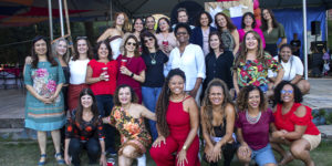 Imagem Acessível: A imagem mostra uma celebração alegre do Núcleo das Mulheres (NM), com destaque para o aniversário de 5 anos do coletivo. Em um espaço ao ar livre com tenda, várias mulheres posam sorridentes para fotos, vestidas com roupas coloridas e alegres, muitas segurando copos. Outras dançam e se divertem ao som do samba. O texto sobreposto diz: “NM comemora aniversário com encontro pra ficar na memória”, destacando o clima festivo e acolhedor do evento. A atmosfera é de confraternização, empoderamento feminino e valorização das conquistas do grupo.