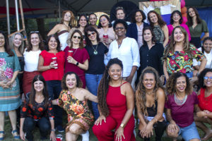 Imagem Acessível: A imagem mostra uma celebração alegre do Núcleo das Mulheres (NM), com destaque para o aniversário de 5 anos do coletivo. Em um espaço ao ar livre com tenda, várias mulheres posam sorridentes para fotos, vestidas com roupas coloridas e alegres, muitas segurando copos. Outras dançam e se divertem ao som do samba. O texto sobreposto diz: “NM comemora aniversário com encontro pra ficar na memória”, destacando o clima festivo e acolhedor do evento. A atmosfera é de confraternização, empoderamento feminino e valorização das conquistas do grupo.