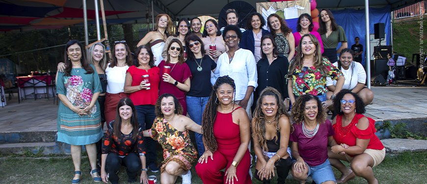 Imagem Acessível: A imagem mostra uma celebração alegre do Núcleo das Mulheres (NM), com destaque para o aniversário de 5 anos do coletivo. Em um espaço ao ar livre com tenda, várias mulheres posam sorridentes para fotos, vestidas com roupas coloridas e alegres, muitas segurando copos. Outras dançam e se divertem ao som do samba. O texto sobreposto diz: “NM comemora aniversário com encontro pra ficar na memória”, destacando o clima festivo e acolhedor do evento. A atmosfera é de confraternização, empoderamento feminino e valorização das conquistas do grupo.