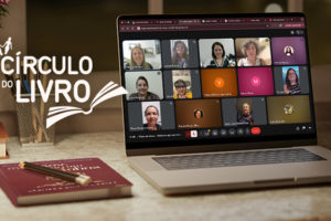 Imagem acessível: A imagem mostra um ambiente aconchegante de estudo e leitura, com luminária, flores e um notebook sobre a mesa exibindo uma reunião virtual com várias mulheres participantes. No topo lê-se “Protagonismo feminino”. O texto principal informa: “NM se reúne para discutir o poder da intuição feminina no Círculo do Livro”. Ao lado aparece o logotipo do projeto Círculo do Livro, indicando o encontro promovido pelo Núcleo das Mulheres do SINJUS-MG para debate e troca de reflexões.