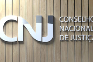 Letreiro em inox do CNJ (Conselho Nacional de Justiça) sobre fundo de madeira. Conteúdo textual: VITÓRIA NO CNJ - FENAJUD GARANTE EXCLUSÃO DE GRUPO PRIORITÁRIO DO CÁLCULO DE 30% DO TELETRABALHO