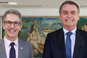 Dois homens brancos vestidos socialmente estão lado a lado e sorriem para a foto. O do lado esquerdo é Romeu Zema, ele tem cabelos grisalhos, olhos castanhos escuros e veste um terno cinza claro. Ao lado dele está Jair Bolsonaro, ele também é grisalho, tem olhos azuis e veste um terno azul marinho.