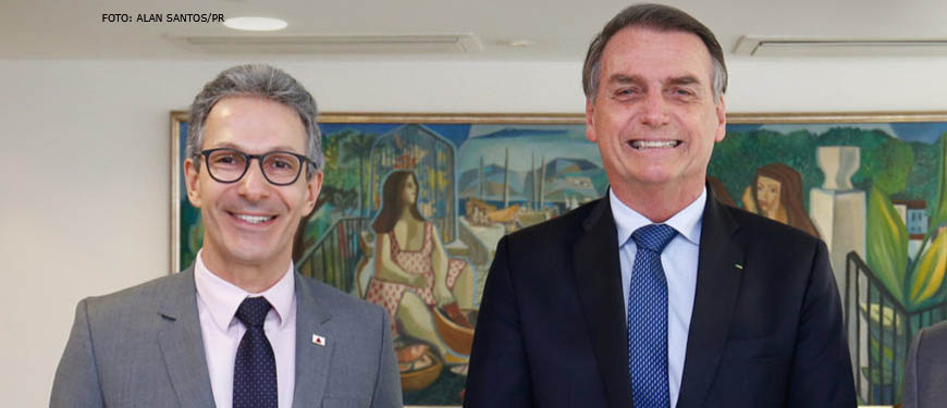 Dois homens brancos vestidos socialmente estão lado a lado e sorriem para a foto. O do lado esquerdo é Romeu Zema, ele tem cabelos grisalhos, olhos castanhos escuros e veste um terno cinza claro. Ao lado dele está Jair Bolsonaro, ele também é grisalho, tem olhos azuis e veste um terno azul marinho.