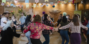 Imagem acessível: A foto ao fundo da imagem representa a comemoração dos 20 anos do Núcleo de Aposentados e Pensionistas (NAP). O registro mostra um amplo salão decorado com diversas bandeirinhas coloridas de festa junina suspensas no teto. No centro, cerca de vinte pessoas do núcleo formam uma grande roda de mãos dadas enquanto dançam. No canto superior esquerdo da imagem está a versal “CELEBRAÇÃO”. Na parte inferior, o texto em distaque diz: “NAP comemora 20 anos em arraiá marcado por forró, dança e união”. Logo abaixo está o símbolo do SINJUS-MG.