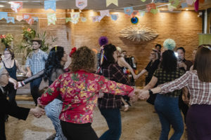 Imagem acessível: A foto ao fundo da imagem representa a comemoração dos 20 anos do Núcleo de Aposentados e Pensionistas (NAP). O registro mostra um amplo salão decorado com diversas bandeirinhas coloridas de festa junina suspensas no teto. No centro, cerca de vinte pessoas do núcleo formam uma grande roda de mãos dadas enquanto dançam. No canto superior esquerdo da imagem está a versal “CELEBRAÇÃO”. Na parte inferior, o texto em distaque diz: “NAP comemora 20 anos em arraiá marcado por forró, dança e união”. Logo abaixo está o símbolo do SINJUS-MG.