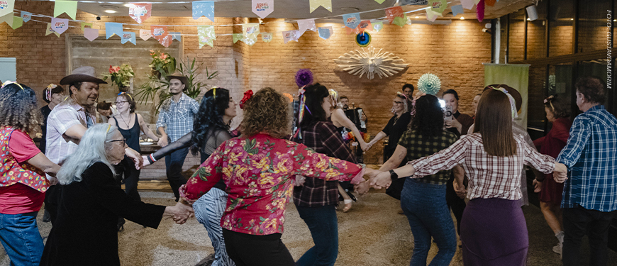 Imagem acessível: A foto ao fundo da imagem representa a comemoração dos 20 anos do Núcleo de Aposentados e Pensionistas (NAP). O registro mostra um amplo salão decorado com diversas bandeirinhas coloridas de festa junina suspensas no teto. No centro, cerca de vinte pessoas do núcleo formam uma grande roda de mãos dadas enquanto dançam. No canto superior esquerdo da imagem está a versal “CELEBRAÇÃO”. Na parte inferior, o texto em distaque diz: “NAP comemora 20 anos em arraiá marcado por forró, dança e união”. Logo abaixo está o símbolo do SINJUS-MG.