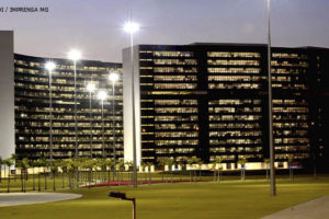 Vista da Cidade Administrativa ao entardecer, dois edifícios curvos, revestidos com vidros escuros em toda a fachada.