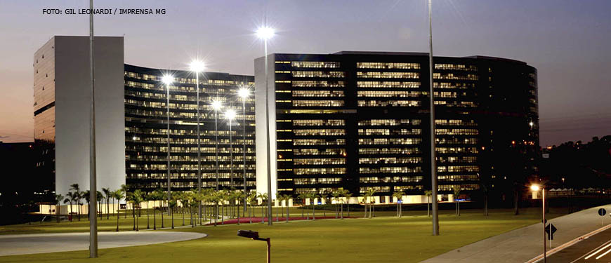 Vista da Cidade Administrativa ao entardecer, dois edifícios curvos, revestidos com vidros escuros em toda a fachada.