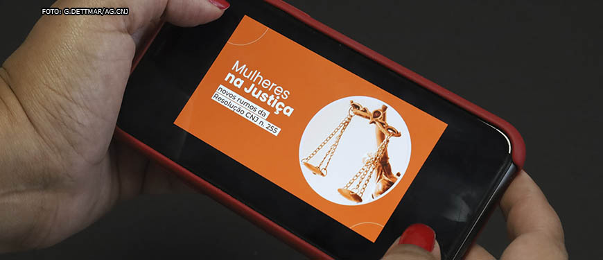 Foto de mãos femininas com unhas pintadas de vermelho segurando um celular na horizontal que mostra o banner com fundo laranja e conteúdo textual " Mulheres na Justiça, novos rumos da Resolução CNJ n. 255", tendo ao lado a imagem de ama balança também em tons de laranja.