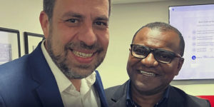 Foto de duas pessoas sorrindo, o ministro da Secretaria Geral da República, Guilherme Boulos e o coordenador-geral da Fenajud, Janivaldo Ribeiro. em ambiente interno, com uma tela ao fundo.