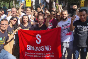 Fotografia com servidores e filiados na luta contra o RRF, na imagem se vê os mesmo com punhos erguidos e a bandeira do SINJUS na cor vermelha.