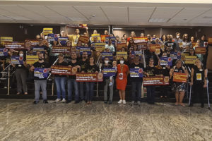 Grupo numeroso de servidores está reunido na escadaria do hall da sede do TJMG, no grupo há mulheres, homens e um bebê de colo acompanhado da mãe. Eles portam cartazes e faixas com palavras de ordem a favor do Teletrabalho.