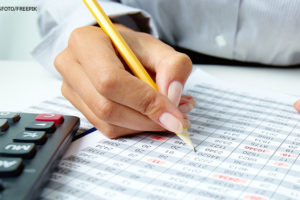 Foto de uma mulher de pele clara em um ambiente de escritório, a mesma segura um lápis amarelo, à sua frente uma folha com diversos números e à esquerda da folha vemos uma calculadora.