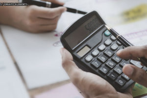 Em destaque estão as mãos de três pessoas em uma mesa cheia de relatórios com gráficos. Duas pessoas manuseiam os papéis e uma delas usa uma calculadora.