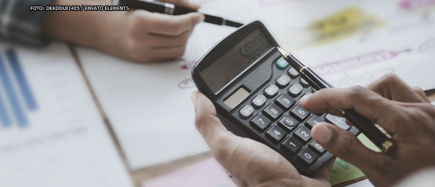 Em destaque estão as mãos de três pessoas em uma mesa cheia de relatórios com gráficos. Duas pessoas manuseiam os papéis e uma delas usa uma calculadora.