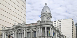 A imagem mostra a fachada imponente da Prefeitura de Juiz de Fora, um edifício histórico de estilo neoclássico, com colunas brancas, janelas altas e uma torre com relógio. Em frente ao prédio, há sinais de trânsito e algumas pessoas passando na calçada. Sobre a imagem, há um destaque em texto que diz: "Feserv Minas reforça que jornada de 30 horas dos servidores de Juiz de Fora deve ser exemplo". Abaixo, o logo da Feserv Minas é exibido com o slogan “Coragem e representação”