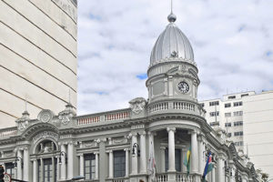 A imagem mostra a fachada imponente da Prefeitura de Juiz de Fora, um edifício histórico de estilo neoclássico, com colunas brancas, janelas altas e uma torre com relógio. Em frente ao prédio, há sinais de trânsito e algumas pessoas passando na calçada. Sobre a imagem, há um destaque em texto que diz: "Feserv Minas reforça que jornada de 30 horas dos servidores de Juiz de Fora deve ser exemplo". Abaixo, o logo da Feserv Minas é exibido com o slogan “Coragem e representação”