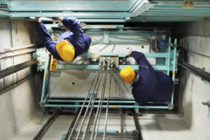 vista aérea de dois homens em um vão de elevador, eles vestem equipamentos de proteção individual e estão realizando a manutenção nas máquinas.