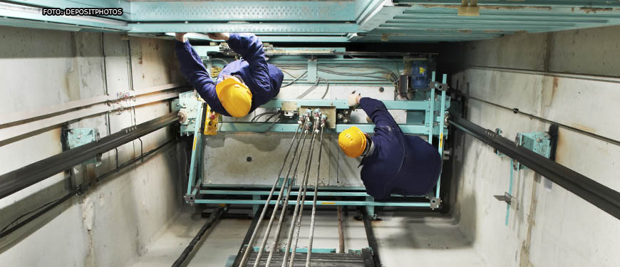 vista aérea de dois homens em um vão de elevador, eles vestem equipamentos de proteção individual e estão realizando a manutenção nas máquinas.