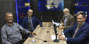 A imagem mostra quatro homens, dentre eles o diretor de Finanças do SINJUS-MG, Alex Aguiar, e o reitor Alessandro Fernandes Moreira, sentados ao redor de uma mesa com microfones, em estúdio com iluminação escura e tela ao fundo com a identidade visual do podcast “Se Liga, Minas”. O texto destaca que o SINJUS debateu os rumos da universidade pública e a inclusão com o reitor da UFMG.