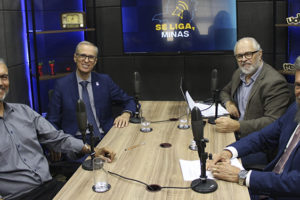 A imagem mostra quatro homens, dentre eles o diretor de Finanças do SINJUS-MG, Alex Aguiar, e o reitor Alessandro Fernandes Moreira, sentados ao redor de uma mesa com microfones, em estúdio com iluminação escura e tela ao fundo com a identidade visual do podcast “Se Liga, Minas”. O texto destaca que o SINJUS debateu os rumos da universidade pública e a inclusão com o reitor da UFMG.