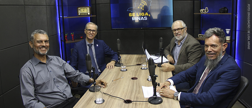 A imagem mostra quatro homens, dentre eles o diretor de Finanças do SINJUS-MG, Alex Aguiar, e o reitor Alessandro Fernandes Moreira, sentados ao redor de uma mesa com microfones, em estúdio com iluminação escura e tela ao fundo com a identidade visual do podcast “Se Liga, Minas”. O texto destaca que o SINJUS debateu os rumos da universidade pública e a inclusão com o reitor da UFMG.