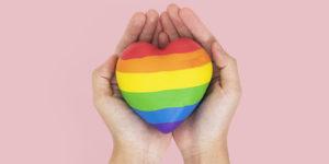 Imagem acessível: A imagem possui fundo rosa e uma foto de duas mãos segurando um coração com as cores da bandeira LGBTQIA+. No canto superior esquerdo, aparece o selo “NOTA DE APOIO”. Em destaque, o texto diz: “SINJUS presta solidariedade à professora vítima de homofobia em escola mineira". No rodapé, há o logotipo do SINJUS.