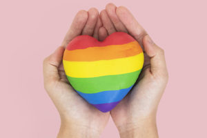 Imagem acessível: A imagem possui fundo rosa e uma foto de duas mãos segurando um coração com as cores da bandeira LGBTQIA+. No canto superior esquerdo, aparece o selo “NOTA DE APOIO”. Em destaque, o texto diz: “SINJUS presta solidariedade à professora vítima de homofobia em escola mineira". No rodapé, há o logotipo do SINJUS.