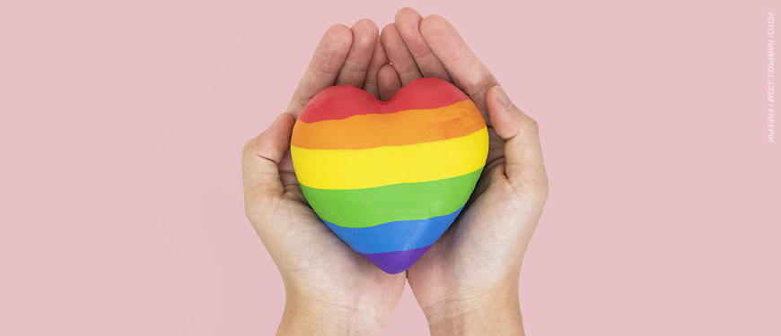 Imagem acessível: A imagem possui fundo rosa e uma foto de duas mãos segurando um coração com as cores da bandeira LGBTQIA+. No canto superior esquerdo, aparece o selo “NOTA DE APOIO”. Em destaque, o texto diz: “SINJUS presta solidariedade à professora vítima de homofobia em escola mineira". No rodapé, há o logotipo do SINJUS.