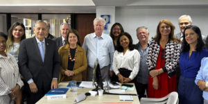 A imagem divulgada pelo SINJUS-MG mostra ao fundo uma foto que registra a atuação da FENAJUD em defesa do fim da contribuição previdenciária de aposentados e pensionistas. A cena mostra uma sala de reuniões com uma mesa retangular ao centro, sobre a qual há documentos, blocos de anotações e placas de identificação de entidades participantes. Ao fundo da mesa, cerca de doze representantes de diferentes entidades sindicais e associativas posam lado a lado para uma foto institucional.