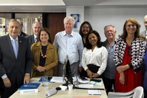 A imagem divulgada pelo SINJUS-MG mostra ao fundo uma foto que registra a atuação da FENAJUD em defesa do fim da contribuição previdenciária de aposentados e pensionistas. A cena mostra uma sala de reuniões com uma mesa retangular ao centro, sobre a qual há documentos, blocos de anotações e placas de identificação de entidades participantes. Ao fundo da mesa, cerca de doze representantes de diferentes entidades sindicais e associativas posam lado a lado para uma foto institucional.