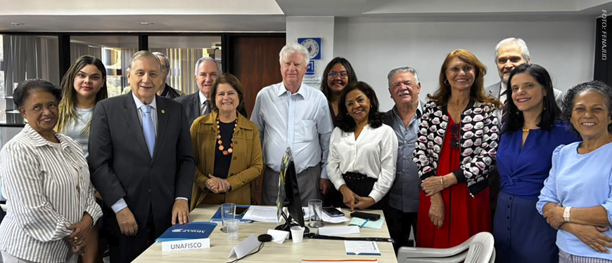 A imagem divulgada pelo SINJUS-MG mostra ao fundo uma foto que registra a atuação da FENAJUD em defesa do fim da contribuição previdenciária de aposentados e pensionistas. A cena mostra uma sala de reuniões com uma mesa retangular ao centro, sobre a qual há documentos, blocos de anotações e placas de identificação de entidades participantes. Ao fundo da mesa, cerca de doze representantes de diferentes entidades sindicais e associativas posam lado a lado para uma foto institucional.