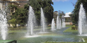 panorama da Praça da Liberdade, ponto turístico da região centro-sul de Belo Horizonte onde há uma concentração de museus e eventos culturais. Em destaque está a fonte luminosa e o coreto, ao fundo está o edifício do CCBB, Centro Cultural Banco do Brasil. Conteúdo textual: Agosto Lilás - PBH cria protocolo para proteger mulheres em espaços de lazer e turismo.